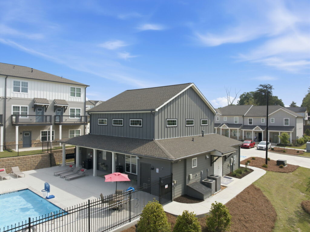 Apartment complex with a pool area in the foreground, gray siding buildings, lounge chairs, and a red umbrella under a blue sky.